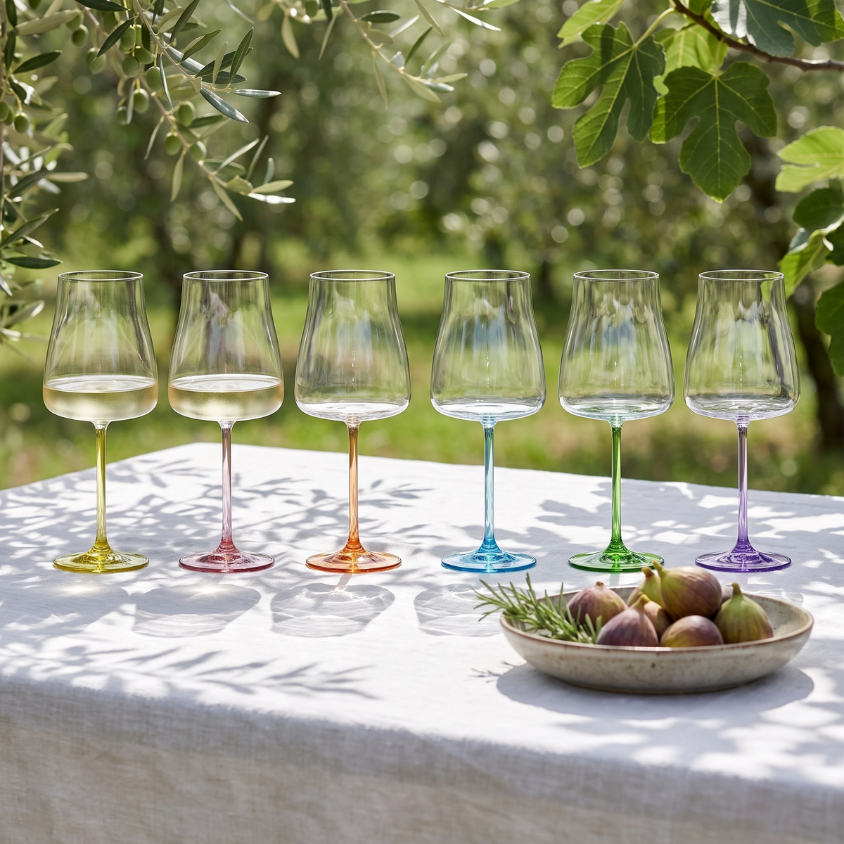 Six Poppy wine glasses with coloured stems on a white linen table in a sunlit olive orchard