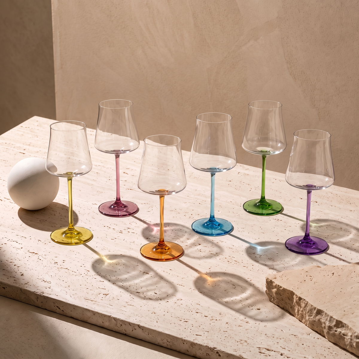 Six Poppy wine glasses on travertine in hard sunlight, coloured stems casting coloured light