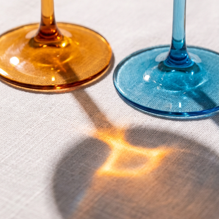 Close-up of amber and blue Poppy wine glass stems and feet casting coloured light on linen